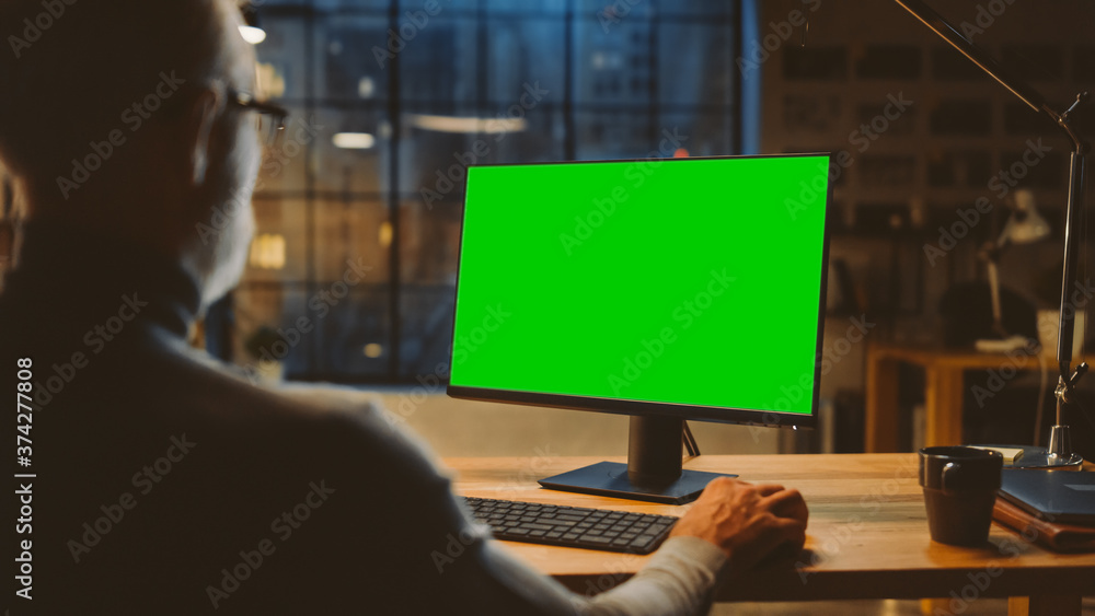 Poster Over the Shoulder: Confident Middle Aged Man Sitting at His Desk Using Desktop Computer with Mock-up Green Screen. Evening in the Stylish Office Studio with City Window View