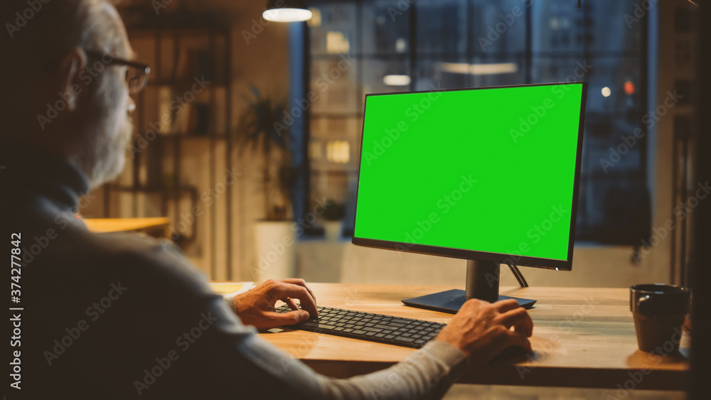 Poster Over the Shoulder: Confident Middle Aged Man Sitting at His Desk Using Desktop Computer with Mock-up Green Screen. Evening in the Stylish Office Studio with City Window View