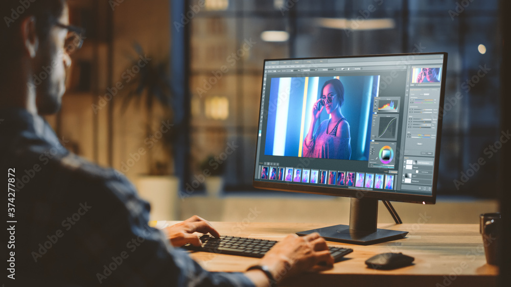 Poster Over the Shoulder: Creative Young Digital Editor Works in Photo Editing Software on His Personal Computer with Big Display. In the Background Evening in Modern Loft Studio Office