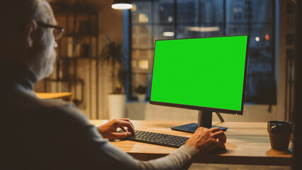 Fototapeta na wymiar Over the Shoulder: Confident Middle Aged Man Sitting at His Desk Using Desktop Computer with Mock-up Green Screen. Evening in the Stylish Office Studio with City Window View