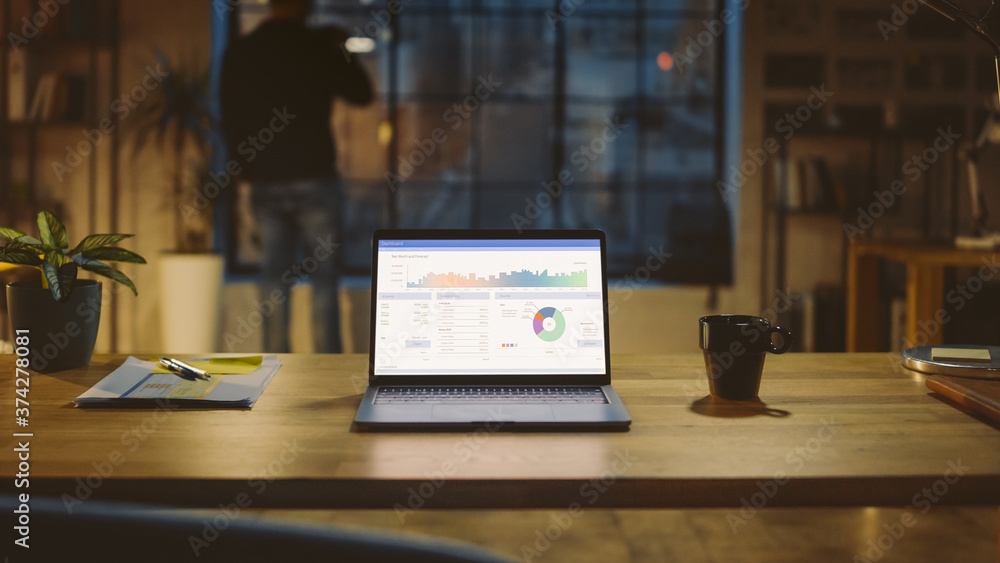 Poster Shot of a Laptop on a Desk with Statistics Graph, Chart and Various Data, Showing Company Growth and Success. In the Background Businessman Drinks from a Cup Looks at the Night City out of the Window