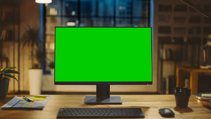 Desktop Computer with Mock-up Green Screen Standing on the Wooden Desk in the Modern Creative Office. In the Background Warm Evening Lighting and Open Space Studio with City Window View. - obrazy, fototapety, plakaty