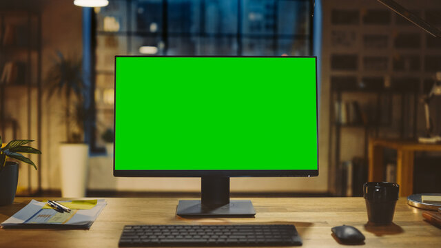 Desktop Computer with Mock-up Green Screen Standing on the Wooden Desk in the Modern Creative Office. In the Background Warm Evening Lighting and Open Space Studio with City Window View.