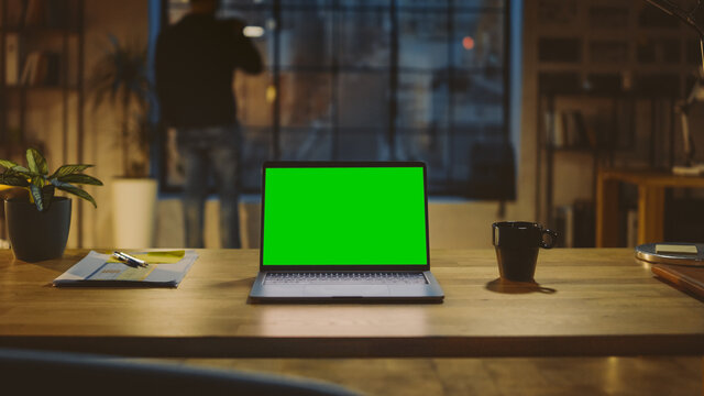 Desktop Computer with Mock-up Green Screen Standing on the Wooden Desk in the Modern Creative Office. In the Background Designer Drinks from a Cup Looks at the Night City out of the Window
