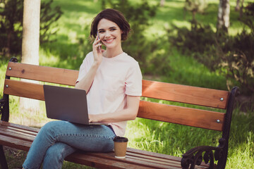 Photo of charming cute lady cheerful look sitting bench hold laptop beaming shiny smiling talk telephone listen family voices weekend abroad wait see them real wear white shirt outdoors