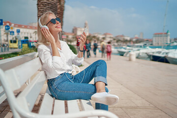Fototapeta na wymiar Enjoying the sound. Happy young woman with earphones listening music on smartphone while sitting on city embankment sea promenade.