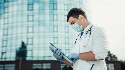 Doctor standing outside with folder full of documents. Man wear medical gown, viral blue face mask, stethoscope and gloves. Coronavirus concept. Image with copy space.