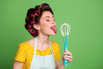 Photo of cute lovely young lady roller hairdo stick out tongue closed eyes licking plastic whisk dough taste before baking wear dotted apron shirt isolated green color background