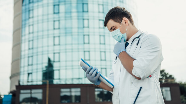 Portrait Of Young Doctor Holding Folder Full Of Documents And Thinking How To Help The Patients. Man Wear Medical Gown, Blue Face Mask, Stethoscope And Gloves. Coronavirus Concept. 