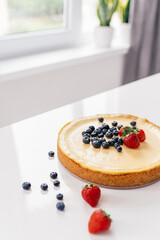 Whole cheesecake with fresh strawberries and blueberries on a white table in white modern kitchen.