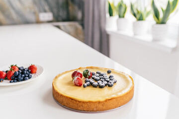 Whole cheesecake with fresh strawberries and blueberries on a white table in white modern kitchen.