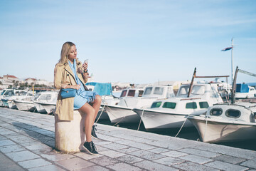 Young traveling woman in coat  sitting on Split promenade sea embankment using smartphone.