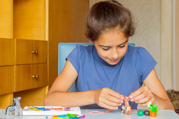 The girl sits at the table at home and sculpts toys from plasticine.