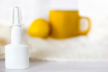 focus on white nasal spray bottle on light background. Yellow cup of tea and lemon in the background. Seasonal disease and runny nose.