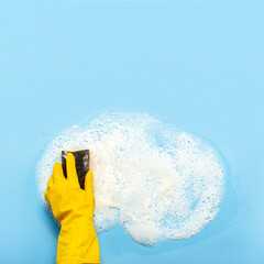 Hand in a yellow rubber glove holds a cleaning sponge and wipes a soapy foam on a blue background....