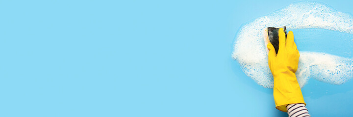 Hand in a yellow rubber glove holds a cleaning sponge and wipes a soapy foam on a blue background....