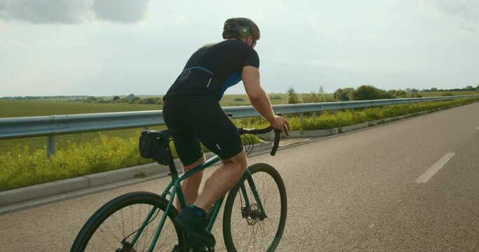 The cyclist is going uphill. He is standing on the pedals and starting spinning them faster. Training on a bicycle. Shooting from behind. Fields in the background. 4K