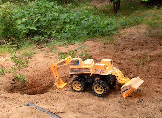 a yellow toy excavator stands on the sand with grass next to a small pit. the picture was taken in a garden in natural light after a rain. bright color.