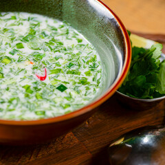Popular spring and summer cold soup okroshka. Okroshka in a bowl, lemon and herbs on rustic wooden table.