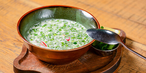 Popular spring and summer cold soup okroshka. Okroshka in a bowl, lemon and herbs on rustic wooden table.