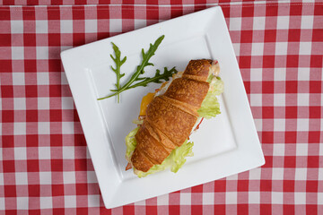 Breakfast with fresh croissants with meat and salad filling served on white plate over red plaid tablecloth.