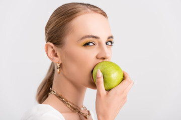 blonde woman biting green apple and looking at camera isolated on white