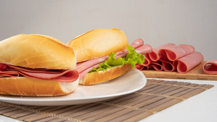 Mortadella sandwich and lettuce on a white plate on a mat and slices of mortadella next to it on a cold board on a table with white tablecloth, selective focus. © Milton Buzon