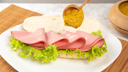 Mortadella and lettuce sandwich, on a white plate and a bamboo mat and pepper sauce and mustard on a table with white towel, selective focus.