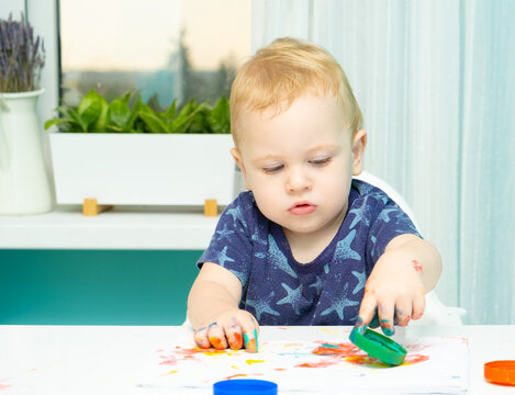 Blond toddler painting with his hands on paper at home using finger paints. Fun activities for toddlers