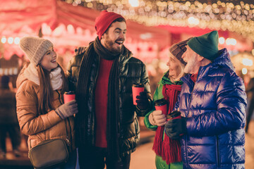 Photo of full family four members x-mas gathering hold mug drink eggnog share news have fun wear outerwear hat scarf coat gloves multi-generation night street park illumination fair outdoors