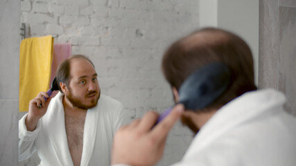 Adult man standing in front of bathroom mirror brushing hair and beard with comb.