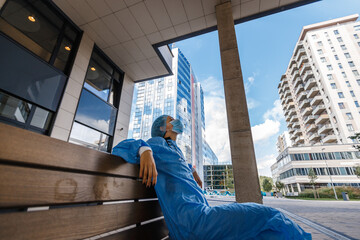 Tired and exhausted doctor takes break on bench near hospital wearing surgical mask, gown and capduring day time