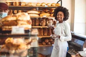 Schilderijen op glas She will have you coming back for more. Gorgeous young woman in her bakery smiling cheerfully to the camera. Smiling young woman selling bread in the bakery. Happy african baker woman © Dragana Gordic