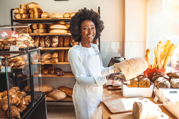 Smiling baker woman standing with fresh bread at bakery. Happy african woman standing in her bake...