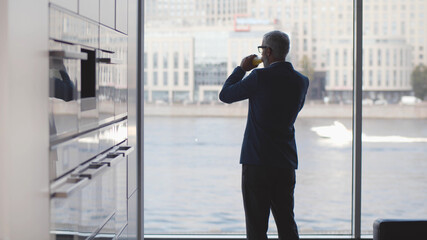 Mature businessman drinking orange juice enjoying window view in kitchen