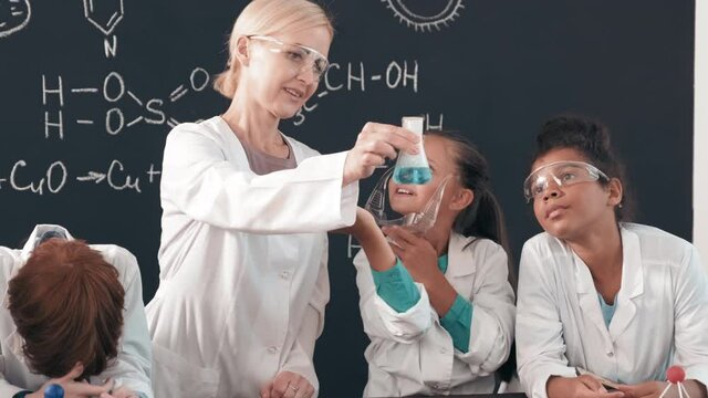 Handheld tracking of female chemistry teacher in lab coat and goggles holding up glass flask with steaming blue liquid and showing it to curious schoolchildren during lesson