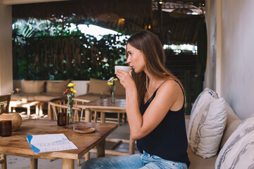 Pensive caucasian female in casual wear resting on break in cafe interior with aroma caffeine beverage, beautiful 20s hipster girl sitting in comfortable cafeteria drinking cappuccino from mug