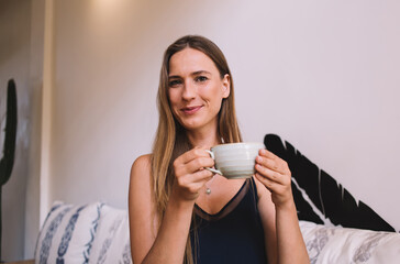 Portrait of smiling caucasian charming female in casual wear holding cup with hot caffeine beverage resting on free time, smiling attractive woman 20s looking at camera enjoying team from ceramic cup