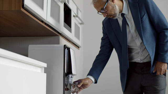 Mature Businessman Getting Water From Water Cooler In Modern Office Kitchen