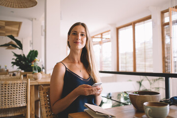Serious caucasian hipster girl in casual wear looking at camera sitting in cafe interior checking messages and mail on cellular, pensive beautiful woman enjoying wireless connection for blogging