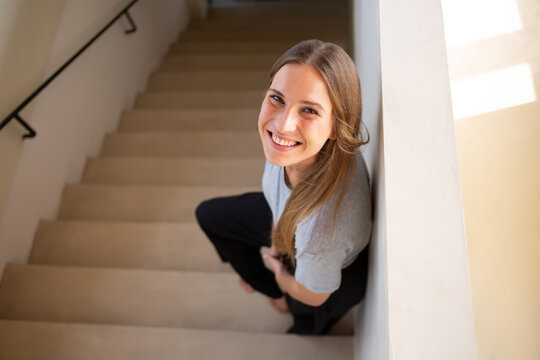 Top view of caucasian happy hipster girl in casual wear standing on stairs enjoying free time smiling looking at camera, from above picture of young 20s with natural beauty skin woman laughing indoors