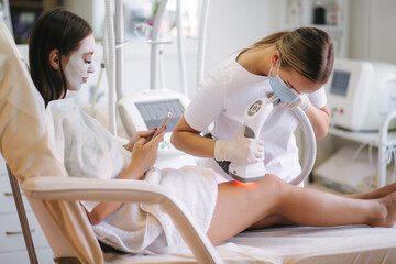 Obraz na płótnie Canvas Beutiful bussines woman with white facial mask using phone while cosmetologist in white gloves performing radiofrequency lifting procedure on the stomach. Beautiful woman in medical clinic. Beautician