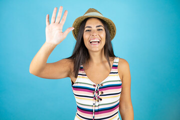 Young beautiful woman wearing swimsuit and hat over isolated blue background smiling and saying hello with her hand up