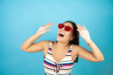 Young beautiful woman wearing swimsuit and sunglasses over isolated blue background scared with her arms up like something falling from above