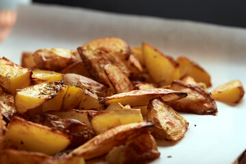 Part of white paper and cutting board with delicious baked golden potatoes slices with seasoning from sideways. On wooden table with free space for text