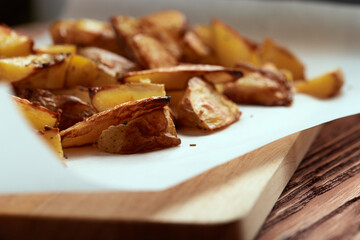 Part of white paper and cutting board with delicious baked golden potatoes slices with seasoning from sideways. On wooden table with free space for text