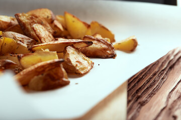 Part of white paper and cutting board with delicious baked golden potatoes slices with seasoning from sideways. On wooden table with free space for text
