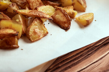 Part of white paper and cutting board with delicious baked golden potatoes slices with seasoning from sideways close up. On wooden table with free space for text