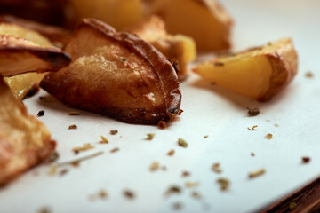 Delicious baked potatoes slices with seasoning on white paper cutting board and wooden table from side close up