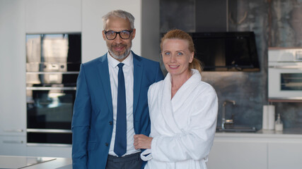 Handsome mature businessman in suit and his wife in bathrobe hugging in home kitchen